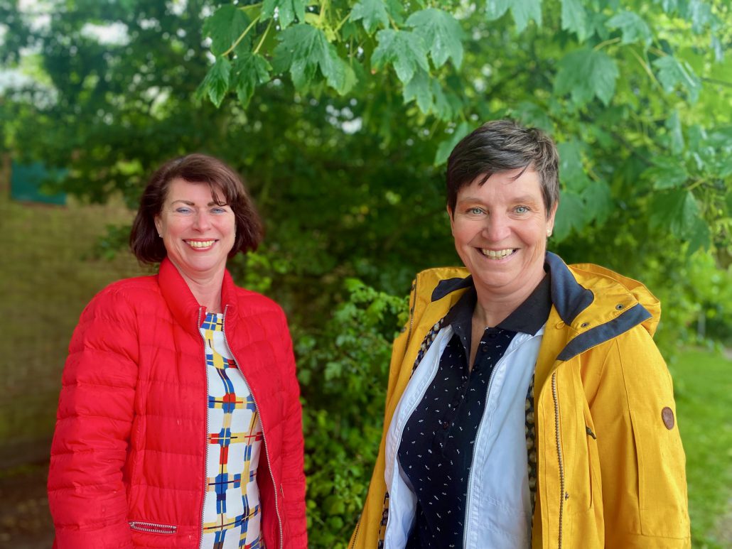Schoolleiders Yvonne de Jager en Monique van Gaalen staan voor een groene boom. Yvonne staat op de voorgrond, glimlacht en draagt een gele jas. Monique staat iets verder naar achteren en draagt een rode jas met een kleurige jurk eronder. Ook zij glimlacht.