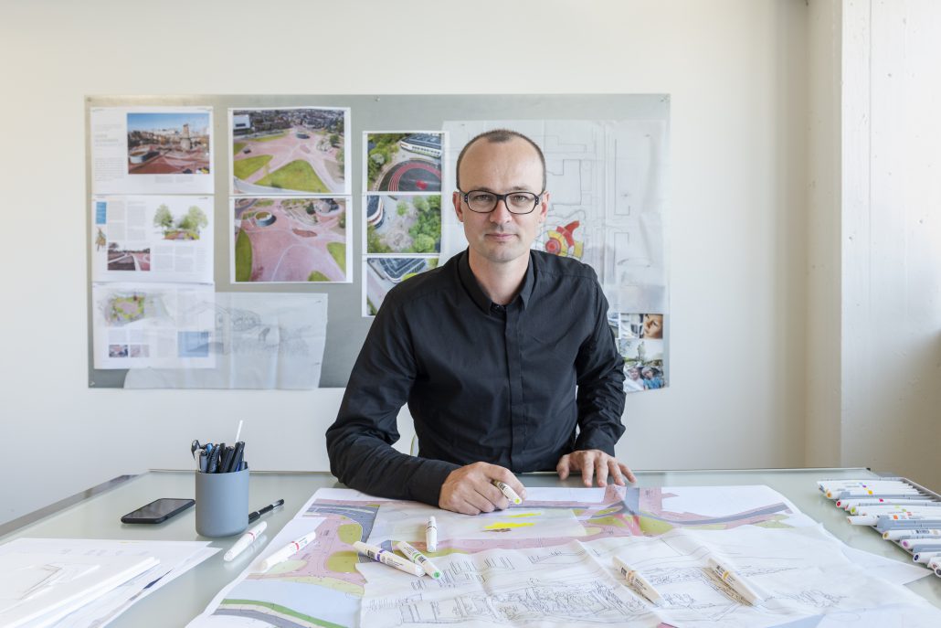 Landschapsarchitect Jeroen Marseille zit achter zijn bureau in een zwarte blouse. Hij draagt een bril. Voor hem op tafel en achter hem aan de wand zijn zijn ontwerpen te zien.