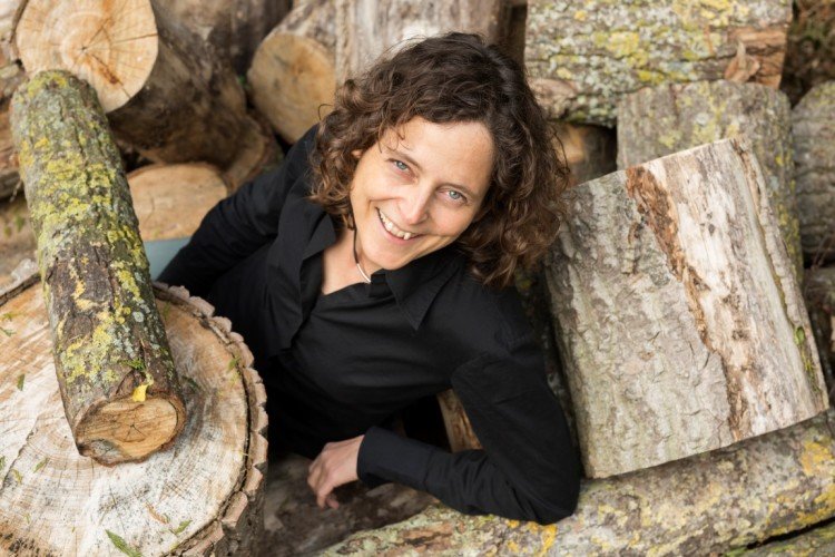 Een vrouw kijkt vrolijk de camera in. Ze zit tussen boomstammen, draagt een zware trui en heeft halflange bruine krullen. De foto is van bovenaf genomen.