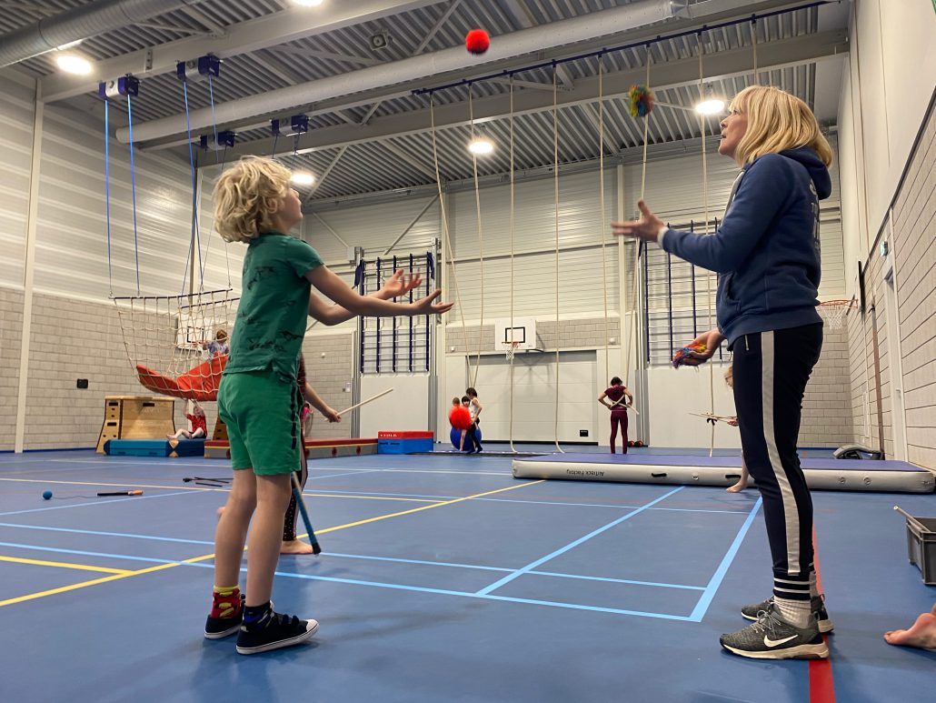 In een gymzaal staat links een blond jongetje in een groen shirt en een groene korte broek te jongleren. Rechts van hem staat een vrouw in een zwarte broek en een donkerblauwe hoodie, ze jongleert ook met een rode bal.