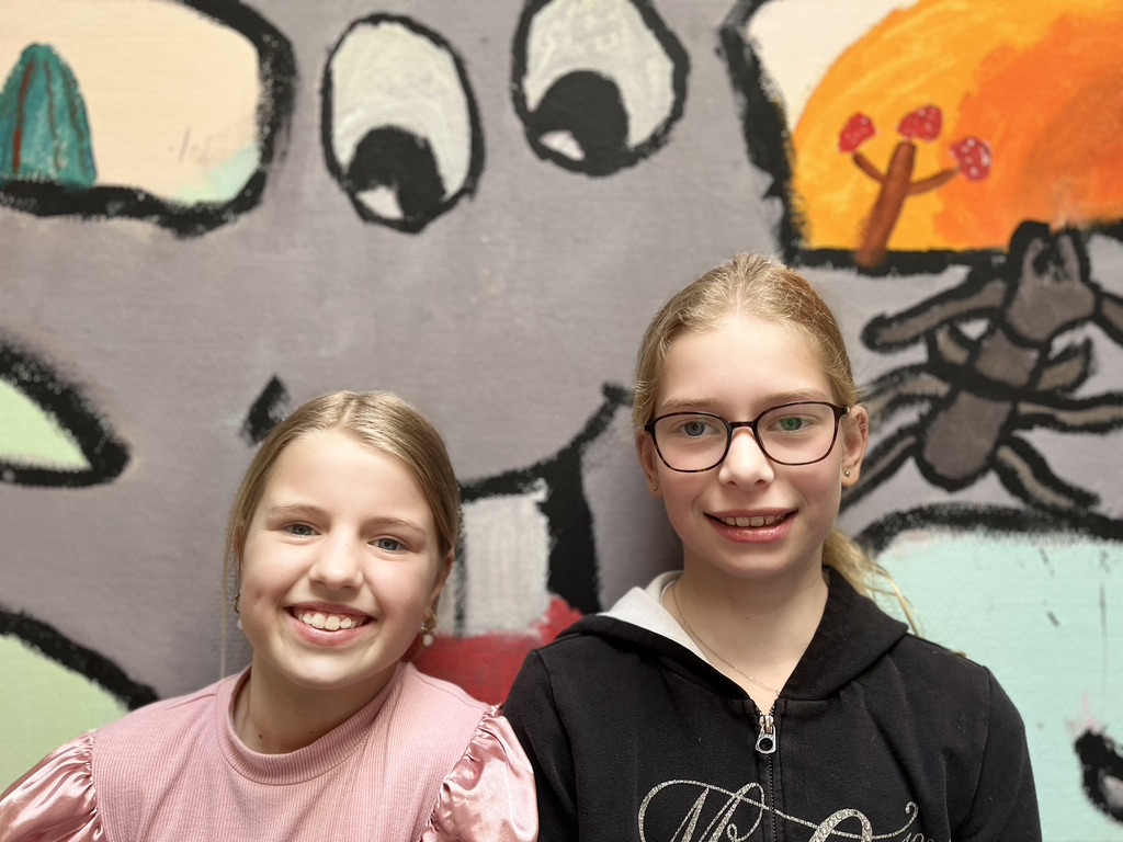 Twee meisjes kijken lachend de camera in tegen een achtergrond waarop een muurschildering staat van een boom. De boom heeft ogen, die boven in het midden te zien zijn.