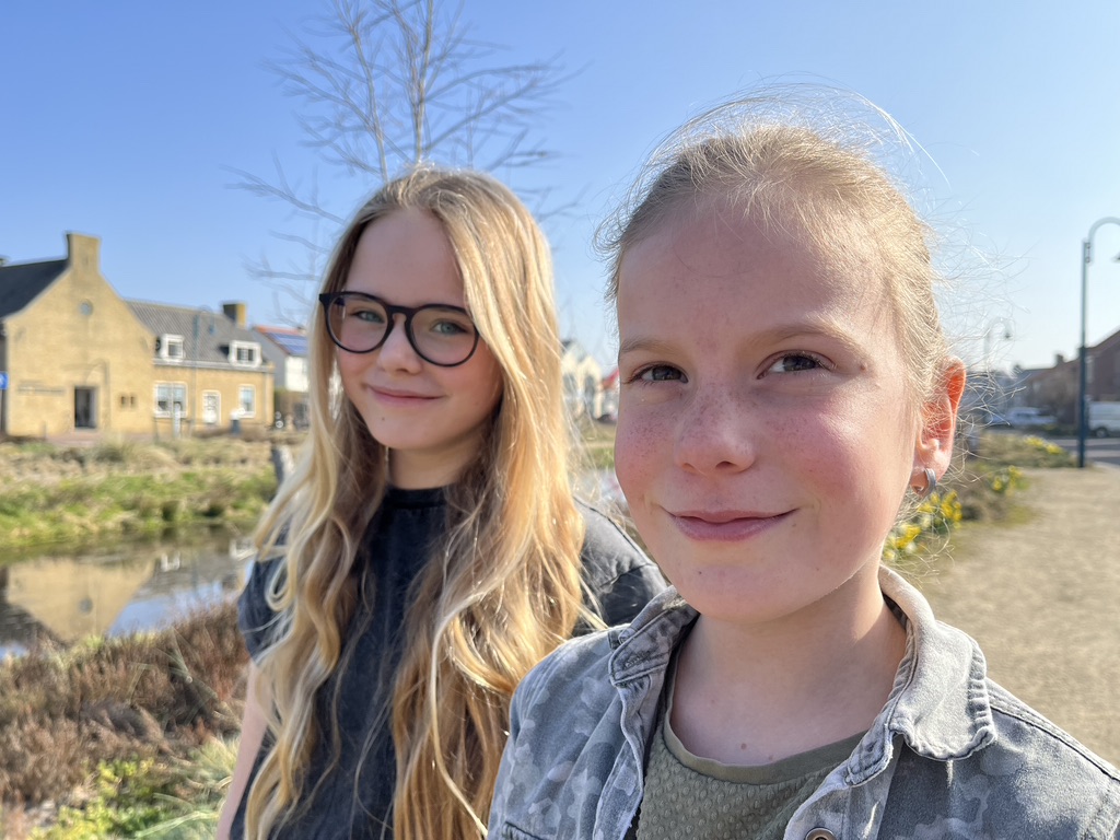 Twee meisjes kijken de camera in met een glimlach op hun gezicht. Ze staan buiten op een zonnige dag. De lucht is blauw en op de achtergrond is een slootje te zien.