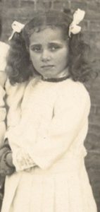 Een meisje met donker haar en staarten kijkt serieus de camera in. De foto is erg oud en stamt van het begin van de 20e eeuw.