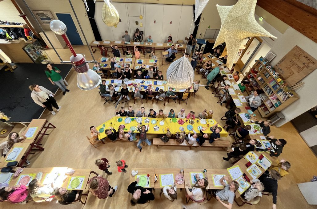 De foto is van bovenaf genomen. Een grote groep kinderen zit aan tafels die gedekt zijn met gele tafelkleden.