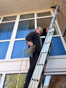 Een man staat op een ladder met een schoolbel in zijn hand. Hij kijkt tevreden.