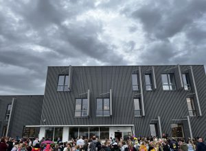 Voor een schoolgebouw staat een grote groep kinderen te wachten met hun ouders. Het gebouw is hoog en van hout.